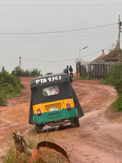 A three-wheeled auto-rickshaw with a black canopy, yellow and green body, and license plate PTA-4965 is parked on a dirt road in Rayfield, Nigeria. Three figures, two men and a woman, stand in the background near a barbed-wire fence and a building with a thatched roof. The environment appears to be rural or semi-urban, with vegetation and overhead power lines visible. The sky is overcast.