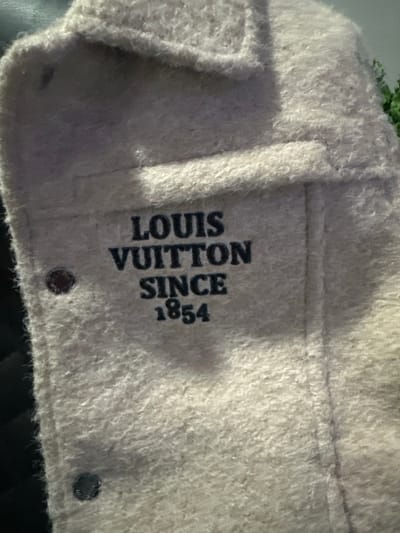 A close-up, low-angle shot shows the exterior of a light-colored fuzzy garment, likely a jacket or coat, with "LOUIS VUITTON SINCE 1854" embroidered in dark block letters across the chest. Two dark, circular buttons are visible, one on the left side of the garment and another lower down. The garment's fabric appears textured and somewhat fuzzy. The background is dark and out of focus. The scene is not depicted within the city of Kazanlak, Bulgaria.