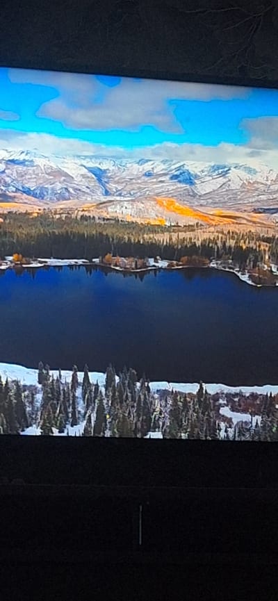 The image depicts an aerial view of a landscape featuring a dark blue lake in the foreground, bordered by a snowy shoreline with evergreen trees. In the middle ground, a dense forest transitions into hills covered in autumnal foliage, with some patches of bright orange. The background is dominated by a mountain range with snow-capped peaks and a bright blue sky with scattered clouds. There is no information about the city of Bila Tserkva, Ukraine, within the provided image.