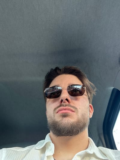 The image is a selfie taken from a low angle, likely inside a vehicle. A man with a beard and sunglasses is the primary subject. He is wearing a white shirt or garment. The background consists of the dark gray interior of the vehicle, with a window visible in the upper part of the frame. The reflection in the sunglasses suggests a cityscape or urban environment outside. While no specific landmark is discernible, the reflections on the sunglasses may be from Glattbrugg, Switzerland.