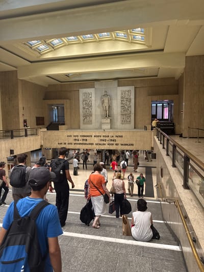 The image shows a group of people inside a large, open building with marble walls and floors. The scene appears to be a public space, possibly a train station or museum hall, given the architecture and the presence of individuals with luggage. The people are of various ages and ethnicities, dressed in casual clothing. Some are standing, while others are seated on stairs or on the floor.
The activity taking place seems to be waiting or gathering, as many individuals are looking around or interacting with each other. The overall atmosphere appears calm and perhaps a bit somber, suggested by the presence of a memorial or monument in the background.
Notable details include the architectural style of the building, with its classical elements and large windows. There is a large inscription on the wall that reads "AUX 3012 CHEMINOTS MORTS POUR LA PATRIE 1914-1918" and "AAN DE 3012 SPOORWEGMANNEN GEVALLEN VOOR HET VADERLAND". This text, written in both French and Dutch, indicates the presence of a war memorial dedicated to railway workers who died for their country. The time of day is not explicitly clear, but the lighting suggests it could be daytime. There is no visible indication of weather conditions as the scene is indoors.
The location cues, specifically the bilingual inscription and the architectural style, strongly suggest that this location is in Brussels, Belgium, a common place for such memorials. The emotion conveyed is primarily one of quiet contemplation or perhaps anticipation, given the context of a memorial site and a public gathering.