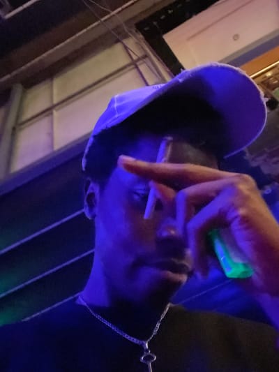 An individual wearing a blue baseball cap and a dark shirt is holding a cigarette to their lips. Their left hand is raised, obscuring part of their face, and they are holding a green object, possibly a lighter, in their right hand. The scene is illuminated by blue and purple lighting, creating a dark, atmospheric environment. The background appears to be indoors, with hints of industrial structures or a dimly lit venue. There is no visible indication of the location being Obosi, Nigeria.