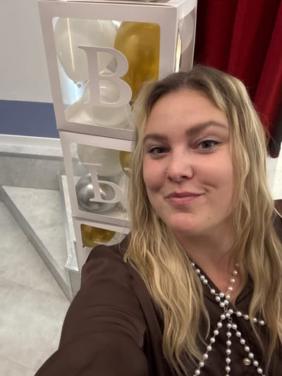 A young woman with blonde hair and light-colored eyes is posing for a selfie. She is wearing a brown satin top and a pearl necklace. In the background, there are white boxes with letters "B" and balloons inside, suggesting a celebration or event. The environment appears to be indoors, with a red curtain visible on the right and a light-colored wall and floor. There is no specific information to indicate the location in Markovskii, Russia.
