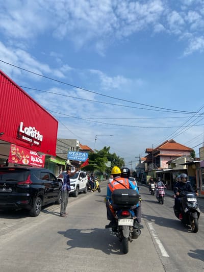 The scene takes place on a street in Kecamatan Tanggulangin, Indonesia, located at latitude -7.5 and longitude 112.702. The street is paved with asphalt and lined with shops and buildings on both sides. The sky is blue with some white clouds. There are several vehicles parked along the street, including a black SUV, a white sedan, and a motorcycle. There are also several people walking and riding motorcycles on the street.  A man in a white shirt and blue jeans is walking on the street. He is waving his left hand in the air. There is a sign that reads "SYAFA' AT. WORN A. JAKET KULIT." Two people are riding a motorcycle down the street. The person in the back is wearing a blue and yellow hard hat. The motorcycle has a black and silver top case.  Several other motorcycles and people are visible in the distance.  The scene is bright and sunny with a few light-colored clouds in the sky.