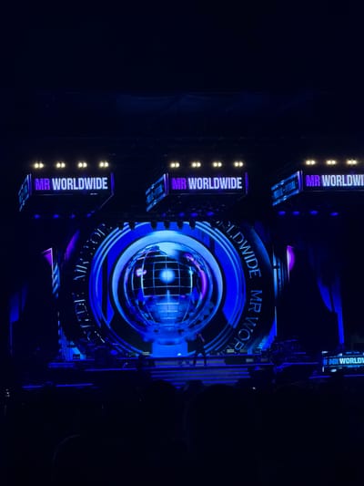 The image depicts a concert stage with a large, circular screen displaying a globe. The stage is illuminated with blue and purple lights. Above the screen, signs read "MR WORLDWIDE." A performer, silhouetted against the light, stands on a raised platform. The audience is visible in the foreground, mostly in shadow. The event appears to be taking place at the Ta' Qali National Stadium in Lija, Malta, suggested by the distinctive architecture visible in the background of some concert photos of the same event.