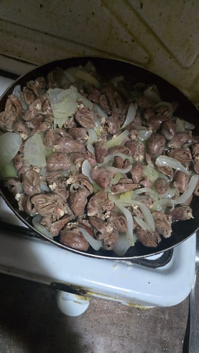 The image shows a close-up, overhead view of a pan filled with cooked chicken hearts and sliced onions, possibly on a stovetop in Kaleytsa, Bulgaria. The pan is dark and appears to be made of metal, with a slightly worn interior. The chicken hearts are small, irregularly shaped, and have a reddish-brown color, suggesting they have been sautéed or stewed. They are mixed with translucent, thinly sliced pieces of white onion. The food fills most of the pan and appears to be the primary subject of the image. The background shows a portion of a white appliance, likely a stove, with some staining or grease marks visible. The lighting is dim, casting shadows and highlighting the textures of the food and the appliance.