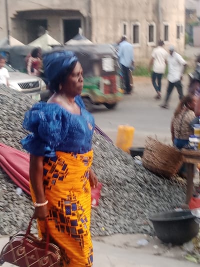 A woman in traditional Nigerian attire, consisting of a blue blouse and a yellow and blue patterned wrapper, stands outdoors carrying a handbag. Her head is wrapped in a blue turban. In the background, a pile of gravel, baskets, and other street vendors' wares are visible. Several people, including men in shirts and trousers, walk along the street behind her. There is also a vehicle with a white front. A building with rectangular windows is in the distance.