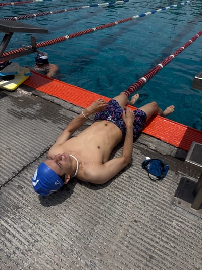 A young man is lying on the concrete poolside of a swimming pool in San Andrés Cholula, Mexico. The man is wearing a blue swim cap, a white beaded necklace, and patterned swim trunks. He is shirtless, and his arms are resting on his stomach. Next to him, a pair of goggles and another swim cap sit. The pool has lane markers, and the side next to the pool is painted orange. The concrete surrounding the pool has a grooved texture. A second person appears in the pool.
