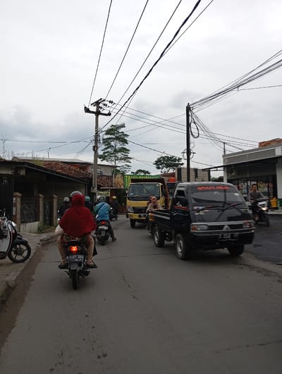 The scene is a narrow, paved street in Kecamatan Kutawaringin, Indonesia. It is daytime, and the sky is overcast. The street is lined with buildings on both sides. Several motorbikes are parked or traveling on the road, one of which is being ridden by a woman wearing a red hijab.  The road is relatively empty, with little traffic.  There are several power lines overhead. A large truck is parked at the end of the road. A few people are visible, including one woman in a red hijab and another woman standing next to the large truck.  Several trees are visible on the left side of the road.