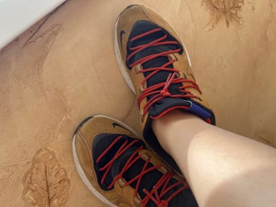 The image shows a person's feet wearing brown and black Nike sneakers with red laces. The sneakers are on a light brown tiled floor with embossed leaf patterns. The scene is lit by natural light, casting shadows. The background is blurred, but appears to be an indoor setting. There is no discernible information in the image to suggest a specific location in Tolyatti, Russia.