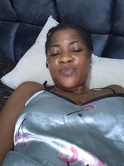 A close-up, eye-level, overhead shot shows a Black woman lying on a white pillow with her eyes closed, seemingly resting or sleeping. She is wearing a light blue satin nightgown with pink floral accents and a thin gold chain with a charm. The background consists of a dark, tufted headboard, typical of a bedroom setting. The lighting appears to be natural, coming from the side. The woman is positioned in the center of the frame. The image was taken in Ajara, Nigeria.