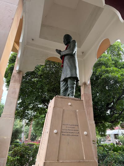 The image depicts a statue of a man in prayer, situated outdoors in what appears to be a park or garden. The statue is made of dark material and is adorned with a red garland. It stands on a tall, tiered pedestal made of light-colored stone, inscribed with text. The surrounding environment includes lush green trees and foliage, with a building visible in the background. The overall scene suggests a memorial or a place of cultural significance. The architectural elements of the structure housing the statue, with its archways and pillars, indicate a deliberate design for public display. The lighting suggests it is daytime, with overcast skies or diffused sunlight contributing to a soft illumination of the scene.
