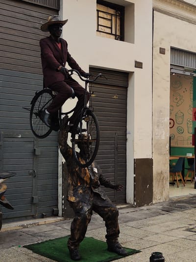 In Lima, Peru, two street performers pose as statues on a city sidewalk. One figure, painted in a dark brown and black, stands on a small patch of green artificial turf, supporting a second performer painted in a similar dark hue, who sits atop a bicycle. The upper performer is clad in a brown suit, a brown hat, and dark gloves, while the supporting performer wears a textured suit of the same color. They are positioned in front of a building with roll-up garage-style doors on the left and the number "808" visible above one doorway. A cafe-style establishment is visible on the right with its interior containing tables and chairs. A jar for tips sits on the sidewalk nearby.