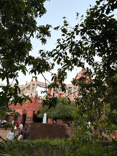 The image captures a view of the ISKCON temple in Delhi, India, seen through a canopy of lush green trees. The temple, constructed with red stone and intricate white architectural elements, features multiple levels and decorative pillars. A prominent dome, with the inscription "ISKCON" visible, stands at the top of the main structure. In the foreground, a fountain with cascading water adds to the serene atmosphere. Several people are gathered around the fountain and on the temple's balconies, engaged in various activities, including walking and conversing. The overall scene depicts a tranquil day at a religious and cultural landmark.
