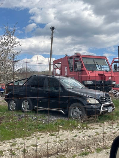 The image shows a black Mercedes-Benz ML-Class SUV parked in front of a red and white fire truck. Both vehicles are located behind a chain-link fence. The background includes a utility pole, a flowering tree, and a cloudy sky. The scene appears to be a junkyard or vehicle storage area. The license plate on the SUV reads "B 6783 BB." There is no indication of people in the image. The vehicles are surrounded by gravel and weeds. The location is assumed to be Resilovo, Bulgaria.