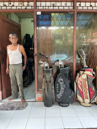 The image shows an elderly Southeast Asian man standing on a tiled floor in what appears to be an outdoor or semi-outdoor setting. He is wearing a white tank top and khaki pants. The man is smiling and looking towards the camera.
Behind him, there are several golf bags and golf clubs. The golf bags are stacked and visible through a window with a diamond-patterned grate. One golf bag is red, white, and black with "TOURSTAGE" written on it. Another is black with "Callaway GOLF" and "BIG BERTHA" visible. A third golf bag is darker in color and shows a crest. The golf clubs are arranged inside the bags, with some shafts and club heads visible.
The setting seems to be a porch or entryway, with a wooden door visible in the lower part of the frame. The window suggests an interior space or a covered area. The lighting suggests it is daytime. The man's relaxed posture and smile convey a sense of contentment. The location cues, such as the tiled floor and the style of the window, might be indicative of a residential setting in Indonesia.