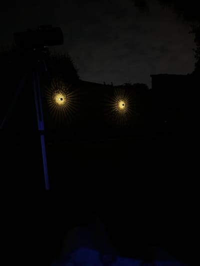 This nighttime image, taken in Villa Pasquali, Italy, depicts an outdoor scene characterized by profound darkness. Two circular, wall-mounted decorative lights are centrally positioned in the mid-ground, each emitting a warm yellow glow and projecting intricate radial light patterns onto the dark wall behind them. To the left, a large, dark-colored telescope or camera mounted on a tripod stands silhouetted against the dark, heavily clouded sky. A vertical blue stripe is visible on one of the telescope's tripod legs. The background reveals indistinct dark outlines of trees and structures. In the extreme bottom foreground, a faint blue illumination is present, obscuring any discernible shapes or objects. No individuals are visible.