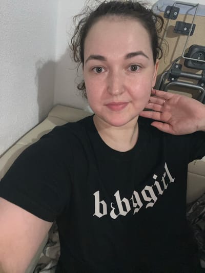 A young woman is shown from the chest up, facing the camera. She has curly brown hair and is wearing a black t-shirt with white gothic lettering that reads "babygirl". Her right hand is raised to her ear, and she is wearing two rings on her fingers and a hoop earring in her visible ear. She appears to be indoors, possibly in a living room, with a neutral-colored sofa or bed behind her. The background also shows a portion of what appears to be a wheeled luggage cart or similar equipment. There is no discernible indication of the city of Chelyabinsk, Russia, or any specific urban location within the image.
