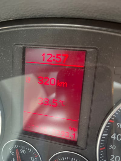 The image displays a close-up of a car's dashboard, showing a digital display with red illuminated text. The display shows the time as 12:57, a fuel level indicator with "H" followed by "320 km", and the temperature as "33.5°C". Below this, the word "trip" is visible, followed by the number "393.1". The surrounding dashboard is dark grey and shows some dust. To the right of the digital display, a portion of a speedometer is visible, with markings for 20, 30, 40, 50, and 90, indicating a speed between 50 and 90. The environment appears to be inside a vehicle. There is no indication of the specific location within Chamboeuf, France, or any people present.