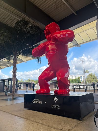 The image displays a large, glossy red, faceted gorilla sculpture by Richard Orlinski, centrally positioned on a black rectangular pedestal. The pedestal's left side features "RICHARD ORLINSKI X", while the right side displays a palm tree logo above "AEROPORT NICE COTE D'AZUR". The sculpture is located outdoors, beneath a corrugated metal roof structure, revealing a bright blue sky with scattered white clouds in the background.
To the left of the sculpture, a tall palm tree with a slender trunk stands prominently. Behind it, a modern bus stop or tram station shelter, partly obscured, shows text indicating "Aeroport Terminal 1". Several individuals are present in the scene: a woman in light jeans and a white top walks near the palm tree on the left; multiple people stand near the bus shelters in the mid-ground, some in dark attire; and two individuals lean on a dark railing in the far right background.
Beyond the pedestrian area and railing, a road is visible with a partially obscured white vehicle, and several street lamps are dispersed. In the distance, white buildings with multiple windows are apparent, one with text partially readable as "Radisson" or similar. The ground is paved with a visible textured pattern. The overall scene depicts an outdoor public space at Nice Côte d'Azur Airport in Nice, France.