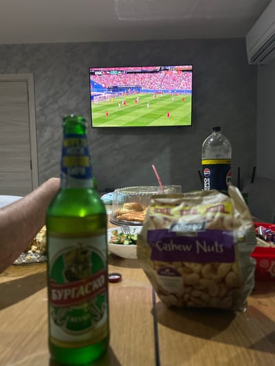 In an indoor setting, likely a living room, a person's hand is visible holding a green glass bottle of Burgasko beer. The bottle's label features Cyrillic script and a crest. In the background, a large television displays a soccer match, with stadium seating filled with spectators. The score on the screen indicates 2-1. Also on a wooden table are a large bag of Alesto Cashew Nuts, a plastic container of food, a clear plastic bottle of Pepsi Zero Sugar, and a red plastic container with items inside. The room has a textured grey wall and a light-colored door on the left. The scene appears to be a casual gathering to watch a sporting event, with snacks and drinks. The location is identified as Belene, Bulgaria, based on the beer label.