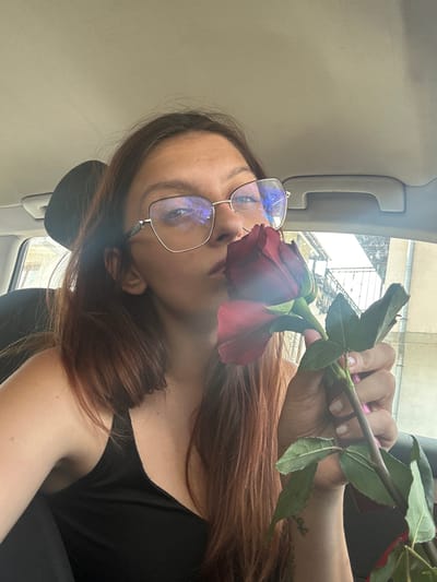 The image shows a young woman sitting in a car, holding a red rose. She is wearing glasses and a black top. The car appears to be parked in an urban environment, with buildings visible through the windows. The woman is leaning towards the rose, as if to smell it, and her eyes are closed. The lighting suggests it is daytime, and the overall atmosphere is relaxed and intimate. While the location is stated to be Tryavna, Bulgaria, there are no specific visual cues in the image to confirm this. The interior of the car is standard, with a headrest and a grab handle visible. The background outside the car is slightly blurred, but hints at a street scene with architectural elements.