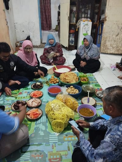 The image shows a gathering of five people in a home in Cangkuang, Indonesia, located at latitude -7.065 and longitude 107.552. Three women are sitting on the floor, wearing headscarves, and one man is sitting on the floor to the right of the image, looking at his phone. A fifth person, a man, is sitting on the floor, facing the viewer, while eating from a plate. The group is gathered around a large spread of food, including a bowl of rice, fried snacks, vegetables, and fruits. The setting is a simple home with a tiled floor. The environment is casual and friendly, suggesting a family gathering or a meal with friends. The image is taken from an angle above the group, capturing a wide view of the scene.