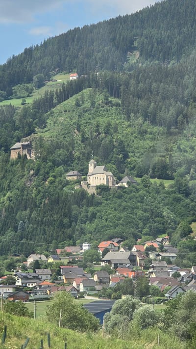 The image shows a panoramic view of the city of Unzmarkt-Frauenburg, Austria, nestled in a valley surrounded by lush green hills covered in dense pine forests. In the upper part of the image, a prominent stone castle, likely Burg Frauenburg, stands on a rocky outcrop, overlooking the valley. Below the castle, a cream-colored church with a tall steeple and clock faces is situated on a hillside. A scattering of residential buildings, varying in color and architectural style, are visible further down the slope, forming a village. In the foreground, a grassy meadow with some scattered trees and a fence suggests a rural setting. The sky above is partly cloudy with patches of blue. The overall scene depicts a tranquil Austrian village integrated into a natural, mountainous landscape.