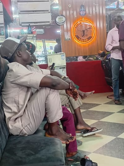 The image displays the interior of KAKNA HAIRCUTTING SALON in Dorayi, Nigeria. In the foreground, a dark-skinned male is seated on a dark grey upholstered sofa, facing right. He wears a light beige long-sleeved shirt, matching trousers, and a beige cap with a mesh back. His bare feet are visible, resting on the sofa, and he holds a dark-colored smartphone in his left hand, with his hands clasped over his knee. Partially visible behind him, another male is seated, wearing dark (possibly burgundy) trousers, with his bare feet resting on the checkered floor next to dark slippers. The floor is tiled with a black and white checkered pattern.
The background reveals a mirrored wall with two white split-unit air conditioners mounted high. A digital clock displaying "4:51" is visible on the wall. Centrally, a circular wooden sign with an orange LED backlight reads "KAKNA HAIRCUTTING SALON," featuring a graphic of the African continent and a barber's pole. To the right, a male stands, wearing a light pink long-sleeved shirt and blue jeans, with sandals on his feet. A red-painted partition or wall section is visible beneath the mirrors on the right side. Reflections of other individuals and salon items are observable in the mirrors.