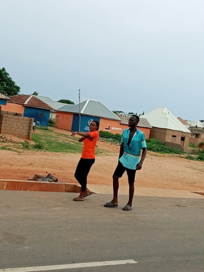 Two individuals, a woman and a man, are standing on an asphalt road in what appears to be an outdoor urban or semi-urban environment in Tumfure, Nigeria. The woman, positioned on the left and facing the viewer, is wearing an orange short-sleeved t-shirt and black leggings. Her arms are crossed at chest level, and she is looking to her left. The man, to her right and facing towards the viewer's left, is wearing an open, short-sleeved teal and patterned shirt and black shorts. He is looking upwards and to his left. Behind them, a landscape of buildings with colorful corrugated metal roofs is visible, some painted orange and blue, and others with natural earth-toned walls. The ground in front of the buildings is sandy and scattered with some debris. The sky above is overcast.