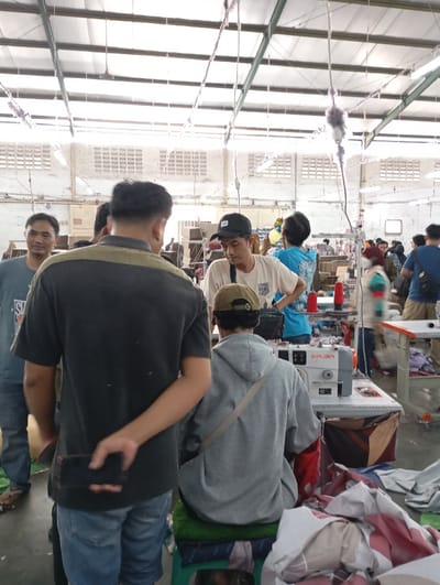 The image captures an active indoor scene in a garment factory or workshop located in Katapang, Indonesia, likely during the daytime. The setting features a high ceiling with exposed metal support beams and corrugated roofing, along with white walls containing multiple ventilation blocks, typical of an industrial building. Wires, cables, and fluorescent lighting fixtures hang from the ceiling.
Numerous individuals, mostly men, are present. In the foreground, a man with his back to the camera, wearing a dark grey t-shirt and blue jeans, holds a black phone behind his back with his right hand. To his left, another man in a blue t-shirt smiles faintly, looking towards the camera. To the right, a man in a light grey hoodie and a cap is seated on a green stool, facing away from the camera, operating a white industrial sewing machine with the brand "SIRUBA" visible on its side. Red and blue spools of thread are on the machine table. Piles of fabric scraps and textile products are scattered on the table next to the sewing machine and on the floor.
Other workers are visible in the mid-ground and background, including a man in a white t-shirt and black cap looking downwards, and another in a blue t-shirt further back. People appear to be engaged in various tasks related to textile production, standing, observing, or actively working at machines. The overall atmosphere suggests a busy, productive environment. No strong emotions are overtly displayed, but the individuals appear focused on their activities.