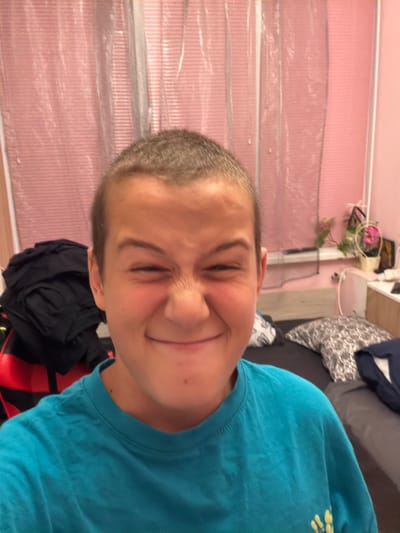 The image shows a close-up, selfie-style photograph of a young person with short, light brown hair. They are making a playful, scrunched-up facial expression, with their eyes squinted and their nose wrinkled. The person is wearing a bright teal t-shirt.
The background appears to be a bedroom. Behind the person, to the left, is a pile of dark clothing. Further back, there is a pink, sheer curtain covering a window or closet. To the right, a bed is visible with pillows and bedding. On a surface behind the bed, there are some plants in pots and other miscellaneous items, suggesting a lived-in space. The overall lighting suggests it is indoors, possibly evening or with artificial lighting.
This scene is likely located in a residential setting, such as a bedroom, within the city of Sliven, Bulgaria. There are no specific landmarks or indicators that pinpoint the exact location within Sliven.