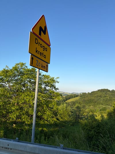 This is a medium-shot, outdoor photograph taken from a roadside perspective. The scene is dominated by a road signpost on the left, with three yellow signs stacked vertically. The uppermost sign is triangular with a red border and depicts a black, zig-zag symbol indicating a winding road ahead. Below this is a rectangular sign with black text that reads "Droga kręta," which translates to "Winding road." The bottommost rectangular sign displays two black arrows pointing upwards, flanking the text "7,0 km," indicating the distance to the end of the winding road.
The road signpost is a metallic grey pole, and to its left, a lush green tree with dense foliage is prominent. The background reveals a rolling, hilly landscape characterized by green fields and dense forests under a clear, bright blue sky. The overall impression is a rural or semi-rural setting, likely in Poland given the Polish text on the sign. No people or animals are visible. The lighting suggests it is daytime, with sunlight casting shadows and illuminating the landscape. A silver metal barrier is visible in the foreground, suggesting the photograph was taken from alongside a road or overlook.