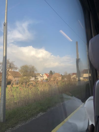 The image depicts a rural-urban transitional landscape in Wilwisheim, France, viewed from inside a moving vehicle, likely a train, looking out a window to the left. The scene is dominated by a clear blue sky with scattered white cumulus clouds. Reflections of the vehicle's interior, including a long rectangular light fixture, are visible on the windowpane.
In the immediate foreground, outside the window, a dark asphalt or paved surface with a distinct yellow line runs parallel to the viewer, edged by white gravel and patches of green grass. Beyond this, a dense field of tall, dried, light brown reeds or grasses extends towards a village. On the far left, a tall, grey metallic pole stands vertically.
The mid-ground features a cluster of residential buildings forming a village. These structures exhibit traditional architecture with stone or stucco facades and varying rooflines, predominantly covered with reddish-brown tiles. A prominent stone building with a dark, conical or pyramidal spire, indicative of a church, is visible on the far left. Deciduous trees, some bare and some with light green foliage, are interspersed among the buildings and fields. A small, distant white vehicle is partially visible within the village.
On the right side of the image, the dark frame of the vehicle's window is visible, along with a partial view of a purple/grey passenger seat and headrest in the reflection. Another tall, grey vertical structure, possibly a railway signal or catenary mast with visible markings, is seen through and reflected on the windowpane. No explicit human figures are discernible in the scene.