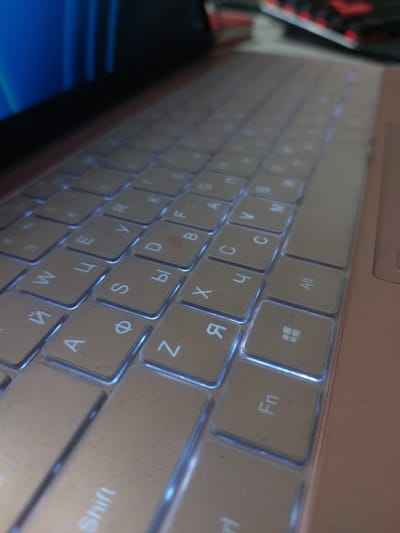 A close-up, high-angle shot shows a rose-gold colored laptop keyboard with illuminated keys. The keys are arranged in rows and are angled towards the top left of the frame. The keyboard appears to be from a device manufactured in Russia, as evidenced by the Cyrillic lettering on some of the keys, such as "Й," "Ф," "Ы," "Я," and "С." The keys are uniformly spaced and have a matte finish. The illumination from the keys casts a blue glow on the surrounding surface. The backdrop is out of focus, showing the corner of a screen with blue and black coloring in the upper left, and a blurred red and black object in the upper right. The overall impression is one of a modern electronic device.
