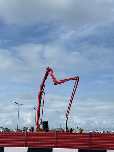 This image captures a daytime construction scene in Igboefon, Nigeria, dominated by a large, red concrete boom pump set against a partly cloudy sky. The photograph is taken from a slightly low angle, looking towards the construction activity.
The central and most prominent object is a bright red concrete pump truck, identifiable by the "SANY" logo visible on its articulating arm. Its multi-jointed boom is fully extended upwards and towards the left side of the frame, poised for or engaged in concrete pouring.
In the mid-ground, a tall red corrugated metal fence topped with several rows of barbed wire runs horizontally across the right half of the image. A black and white striped barrier or post is positioned at the far right edge of the fence. Behind this fence, several construction workers are visible. They are wearing standard personal protective equipment, including high-visibility vests (some yellow, some green) and hard hats, suggesting they are actively involved in supervising or performing tasks related to the concrete operation. A black tank, possibly for water or fuel, and the cab of another construction vehicle (likely a concrete mixer or support truck) are also partially visible behind the fence.
In the lower-left quadrant, a modern street light or utility pole with a dark, flat head stands against the sky. The sky itself is a bright blue, interspersed with scattered white cumulus and cirrus clouds, indicating fair weather conditions suitable for outdoor work. The overall impression is one of active industrial construction taking place under clear daylight.
