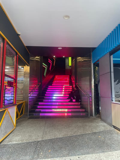 In Surfers Paradise, Australia, an exterior view shows a bright, modern entrance to a venue. The main focal point is a wide staircase illuminated with vibrant pink and purple LED lights on each step. The stairs are bordered by sleek, metallic walls that reflect the colorful light. On the left side of the image, a section of a building facade features a red and black exterior with a yellow trim. A digital display board, showing images of people and text, is visible. A security camera is mounted on the dark ceiling above this entrance. A lone individual, dressed in dark clothing, is seated on the illuminated staircase, looking down and holding a phone. Above the staircase, neon lights in the shape of arrows, in green and pink, are mounted on the dark ceiling. To the right of the staircase, a portion of a building facade with blue vertical slats is visible, and a large, stylized black and white graphic of a face is partially seen on the wall. The ground in front of the entrance is paved with speckled concrete.