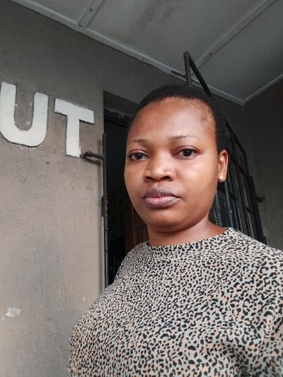 A medium close-up photograph captures an adult female, facing forward with a neutral expression and direct gaze towards the camera. She possesses a dark complexion, short dark hair, and wears a light-colored top patterned with dark leopard spots. A small, round, gold-toned earring is visible in her right ear. To her left, the background consists of a light grey-brown textured wall featuring two prominent, raised white capital letters, "UT". To her right and slightly behind her, a dark recessed area, consistent with a doorway or window, is partially visible, obscured by dark metal bars. The upper frame reveals an off-white soffit or ceiling structure with visible paneling, from which a dark metal rod protrudes vertically on the right side. The scene depicts an exterior or semi-enclosed architectural space in the city of Agani, Nigeria.
