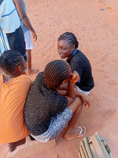 Three girls are resting on a sandy surface. One girl, wearing a black patterned shirt and checkered shorts, sits behind another girl in an orange shirt. A third girl, wearing a black and white striped shirt, rests on the sand with one arm outstretched. One girl wears a black shirt with blue trim and has braided hair. Sand and bags are visible.