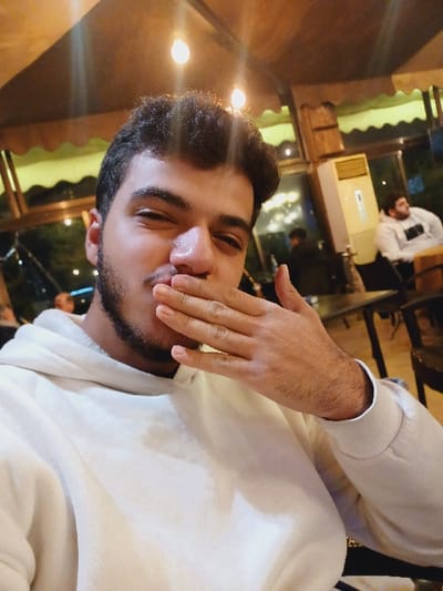 A person with short, dark hair and a trimmed beard is facing the camera, partially covering their mouth with their hand. The individual is wearing a white hooded sweatshirt. The scene appears to be indoors, possibly a restaurant, with soft lighting. In the background, other people can be seen, along with window reflections and restaurant decor. The image is slightly tilted, adding a casual feel.