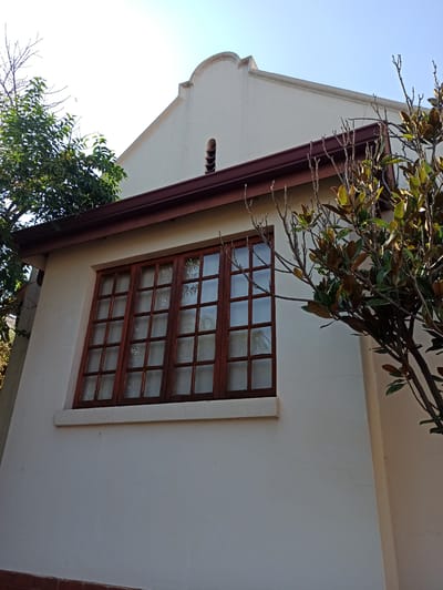 The image shows a close-up view of the exterior of a white building in Pretoria, South Africa. The building has a light beige-colored gabled roof with a dark brown gutter system. A large wooden window with multiple panes is visible on the side of the building. The window has a light-colored sill. Trees with green leaves and bare branches are present in front of and to the side of the building. The sky is clear and blue. The photo appears to have been taken during the daytime. The building has a simple architectural style with a decorative element at the peak of the gable. There is a small, round vent visible on the gable. The wall of the building has some minor cracks.