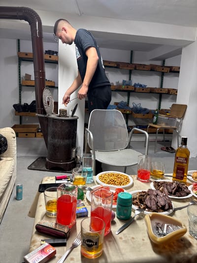 An adult male stands to the left of a wood-burning stove with a stovepipe, apparently stoking it, in a room that appears to be located in Zetiovo, Bulgaria. The room has shelving on the walls filled with various items. A table laden with food and drinks, including a bottle of "Hankey Bannister" whiskey, several glasses of beverages, plates of meat and nuts, and an ashtray filled with cigarette butts, sits in the center of the room. A metal chair is tucked under the table. A portion of a light-colored sofa is visible in the foreground.