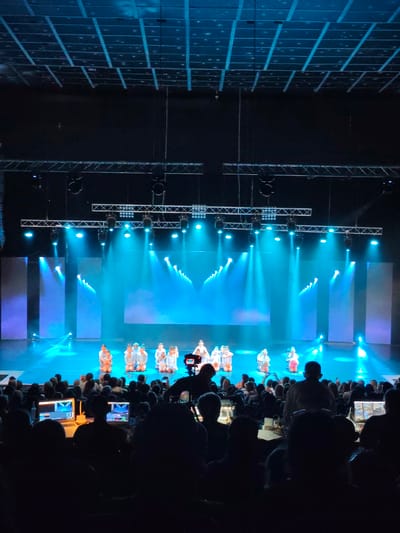 This image depicts a live performance on a stage with a large audience in the foreground. The stage is illuminated with blue and white lights, creating a dramatic effect. Several children, dressed in light-colored costumes, are positioned on the stage, some kneeling and others standing. The background of the stage features large screens displaying abstract patterns of light. Above the stage, a complex rigging system with lights and sound equipment is visible.

In the audience area, many people are seated in the dark, with some individuals in the front rows engaged with laptops and other electronic devices, suggesting they might be operating technical equipment for the performance. The overall atmosphere suggests an indoor event, possibly a dance recital or a theater production.

The description provided does not include information about the specific location in the city of Mykolaiv, Ukraine, as that detail is not present in the image itself.