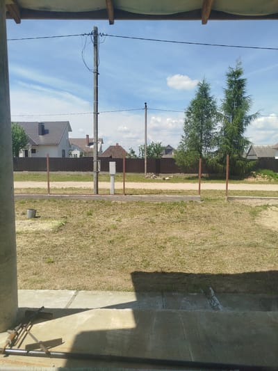 The image depicts an outdoor scene under a partly cloudy blue sky, with a focus on a residential area in Крывасельскі сельскі Савет, Belarus. In the foreground, a concrete structure casts a shadow across a concrete surface where several metal tools are placed. Behind this, a strip of dry, mowed grass is visible, bordered by a concrete foundation and a wire fence supported by metal posts. Beyond the fence, a dirt road runs parallel to a dark brown corrugated metal fence, behind which several houses with tiled roofs are situated. Two tall, green coniferous trees stand prominently in the middle ground. A concrete utility pole with electrical wires and a white utility box are also visible. In the distance, more houses and trees can be seen under the sky. The overall impression is one of a rural or suburban residential environment on a clear day.