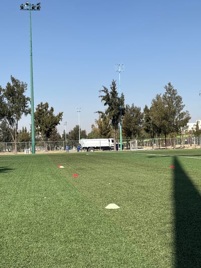 The scene depicts an artificial turf sports field located in a sports complex in León, Mexico, under a clear blue sky. In the foreground, the green artificial turf is marked by four disc cones: one light green and three red. A long, dark shadow extends from the lower right corner towards the center of the field. In the midground, a chain-link fence separates the playing surface from an asphalt or paved service area.
A white service truck, appearing to be a tanker or utility vehicle, is parked parallel to the fence on the paved area. Two individuals are present near the truck; one, dressed in a blue uniform, stands to the left side of the truck, while another individual, also in work attire, is partially visible to the right of the truck. Their actions suggest attendance to the vehicle or the immediate surroundings.
Beyond the fence and truck, multiple tall poles rise against the sky. On the far left, a prominent green pole equipped with several floodlights at its apex is visible. To the right, another tall, slender pole, painted blue and topped with a square framework, stands amongst a line of dense green trees that form the background. Additional blue-painted light poles are discernible in the distance, further integrating the scene into a structured outdoor recreational facility. A light-colored building is faintly visible in the far right background, behind the trees.