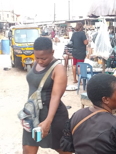 The image captures a vibrant and busy market scene in what appears to be Akpaka Forest Reser, Nigeria. In the immediate foreground, a woman walks towards the left, her gaze directed downwards. She wears a dark grey or black sleeveless dress and carries a light-colored shoulder bag featuring a Union Jack design on one pocket. In her right hand, she holds a light blue object, possibly a wallet or phone, along with another dark item. To her right, the back of another person's head and shoulder is visible, wearing a black top with lace detailing on the sleeve and a brown bag strap across their shoulder.
The midground features a yellow motorized rickshaw, commonly known as a Keke NAPEP, with the words "GOD CALL" and "TVS" visible on its front. Several other individuals populate the scene; one person in a black shirt and red shorts stands facing away, while another in a striped shirt sits on a stool. A blue plastic chair is placed on the ground nearby. The background reveals a bustling market environment with numerous stalls, temporary structures, and more people, suggesting active commerce. The ground is a mix of dirt and sand. The bright, diffused lighting indicates it is daytime, possibly under an overcast sky or bright sun, without casting harsh shadows. The overall impression is one of a functional and lively outdoor market.