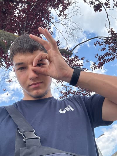 The media file shows a close-up, low-angle shot of a young person looking up at the sky, with a hand held up to their face, obscuring part of it. The subject is wearing a dark blue t-shirt with a white logo on it and a black watch on their left wrist. The background is dominated by a blue sky with white clouds, and the branches of a tree with dark red leaves. The perspective suggests the photo was taken outdoors, possibly in a park or garden. The lighting indicates it is daytime, likely afternoon given the brightness. The overall mood is casual and playful. There is a strap with a buckle visible on the right side of the frame, which could be part of a bag or backpack. The logo on the t-shirt is difficult to discern clearly, but appears to be a stylized design. No other text is visible in the image. The location context is Mannheim, Germany.