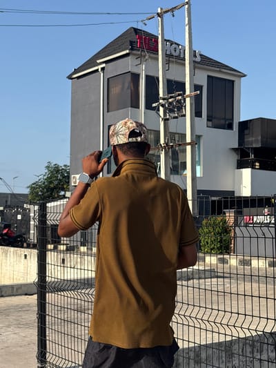 In Igboefon, Nigeria, a person is observed from behind, standing outdoors and holding a dark-colored smartphone to their right ear, appearing to be on a call. The individual wears a patterned baseball cap, a ribbed olive-brown short-sleeved collared shirt with dark trim on the sleeves, and dark shorts. A silver-toned smartwatch is visible on their left wrist. In the background, a multi-story building with grey walls, dark tinted windows, and a dark pitched roof is prominently featured. Red and white illuminated signage on the building's upper facade and roofline spells "TIZ HOTELS." A concrete utility pole, equipped with multiple horizontal cross arms, insulators, and electrical wiring, stands in the midground between the person and the hotel building. A black wire mesh fence occupies the immediate foreground. The ground surface is paved, and the sky overhead is clear and blue. Some green foliage is visible near the base of the hotel and behind the fence. A dark-colored motorcycle or scooter is partially visible parked behind the fence to the far left.