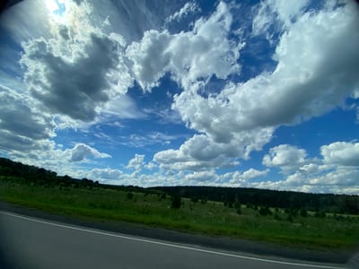 A wide-angle, low-angle shot captures a bright blue sky filled with dramatic, puffy white and gray clouds, some of which are illuminated by the sun, creating lens flare at the top left of the frame. The clouds are varied in size and density, with some appearing wispy and others thick and voluminous. Below the sky, a green, rolling landscape stretches horizontally across the frame. This landscape is covered in grass, with scattered trees and bushes, and a dense, dark green forest line forming the horizon. In the foreground, a portion of an asphalt road with a white line is visible on the left side, suggesting the viewpoint is from a vehicle traveling along it. The scene appears to be a rural or natural setting, possibly in the city of Tyulyugul, Russia, on a day with dynamic cloud cover. There are no people or animals visible in the image.