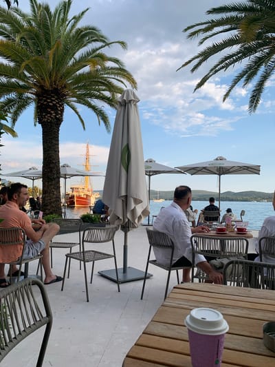 The image depicts an outdoor seating area of a cafe in Tivat, Montenegro, with a view of the Adriatic Sea. In the foreground, several tables and chairs are arranged on a tiled patio. A large palm tree dominates the left side of the frame, its fronds reaching towards a partly cloudy sky. In the background, the sea is visible, with a large, red and wooden sailing ship anchored in the distance. Several people are seated at tables, some facing the water, others engaged in conversation. One person in the foreground is wearing an orange shirt and shorts, seated at a table with a drink. Another individual, in a white shirt, is seated at a table with two red cups and saucers. The overall atmosphere suggests a relaxed dining or drinking experience by the waterfront.